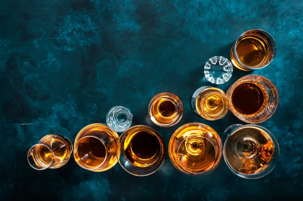 An overhead view of assorted glasses of liquor on a dark teal background, representing the variety of drinks available in liquor shops and bars in Singapore .